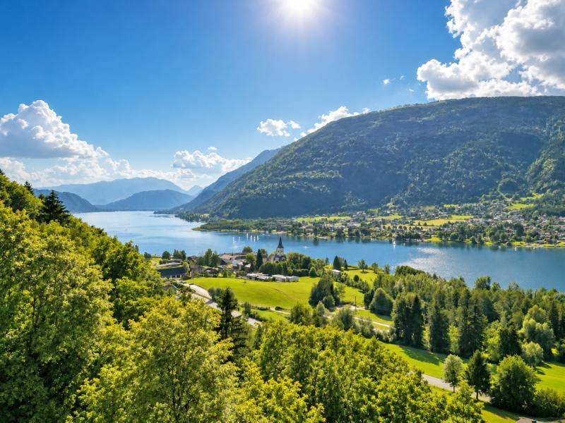 Panoramablick auf den See, umgeben von grünen Hügeln und Wäldern unter blauem Himmel.