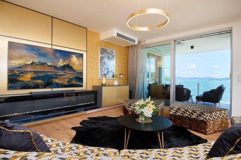 Modernes Wohnzimmer mit großem Fernseher, stilvollen Möbeln und Zugang zur Terrasse mit Seeblick.