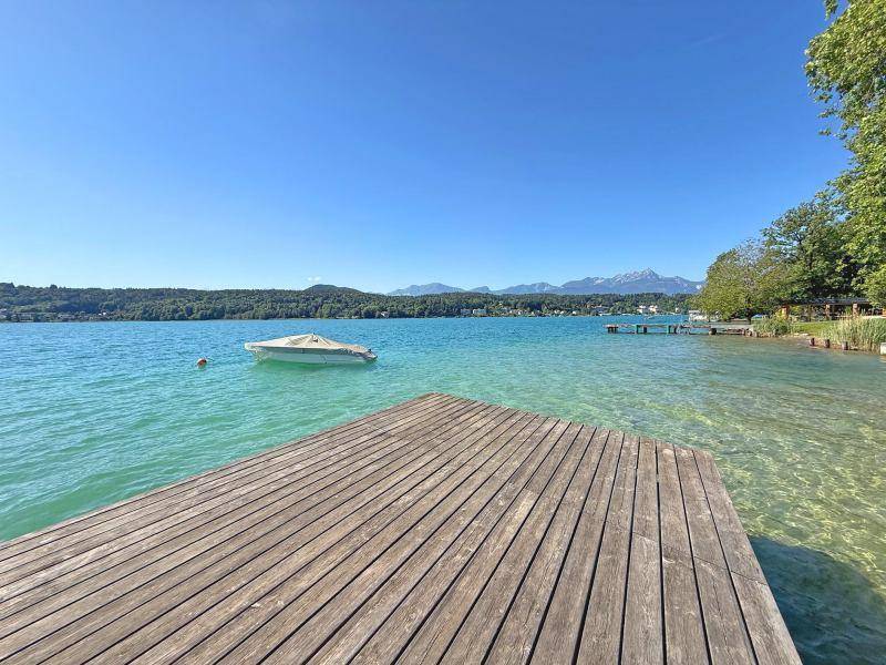 Langer Holzsteg ragt in den türkisfarbenen See, mit einem Boot und Bergen am Horizont.