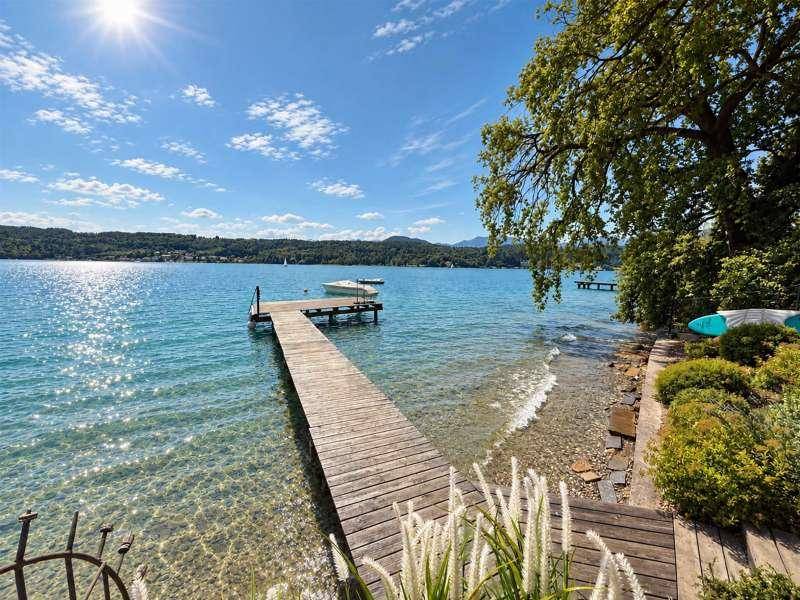 Sonniger Seezugang mit Holzsteg und klarem Wasser, umgeben von grüner Natur und Bergen.