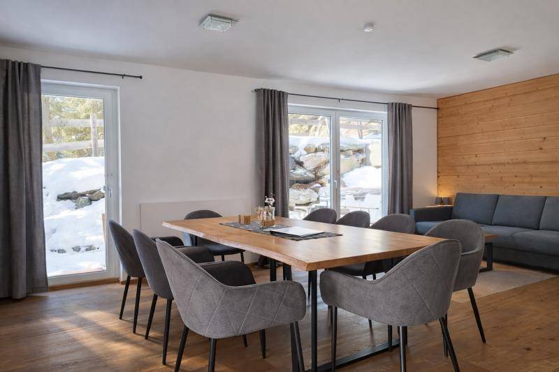 Moderner Wohn-Essbereich mit Holztisch, grauen Stühlen und Sofa, umgeben von großen Fenstern mit Winterblick.
