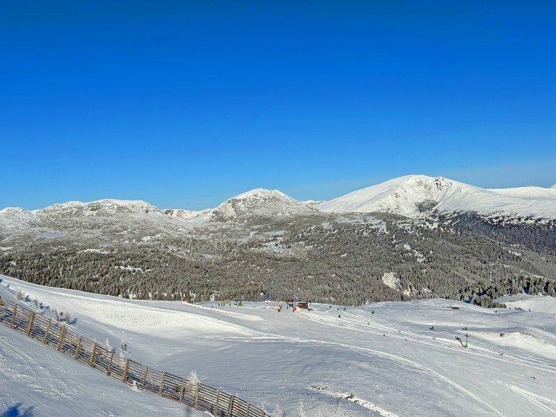 Breite, sonnige Skipiste mit einer Holzzaunbegrenzung und einem weiten Blick auf die schneebedeckten Gipfel der Alpen.