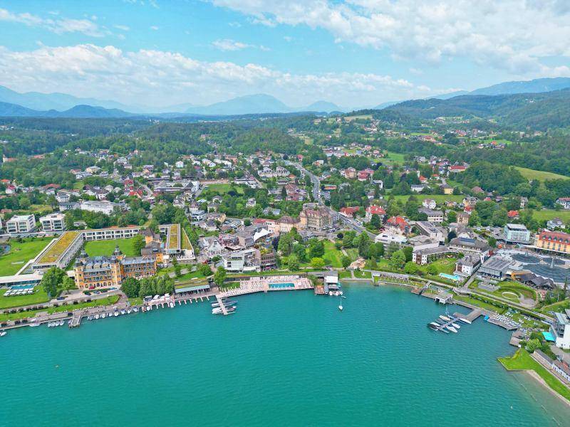 Panoramablick auf eine Stadt am Seeufer mit Gebäuden, Bootsanlegestellen und Bergen im Hintergrund.