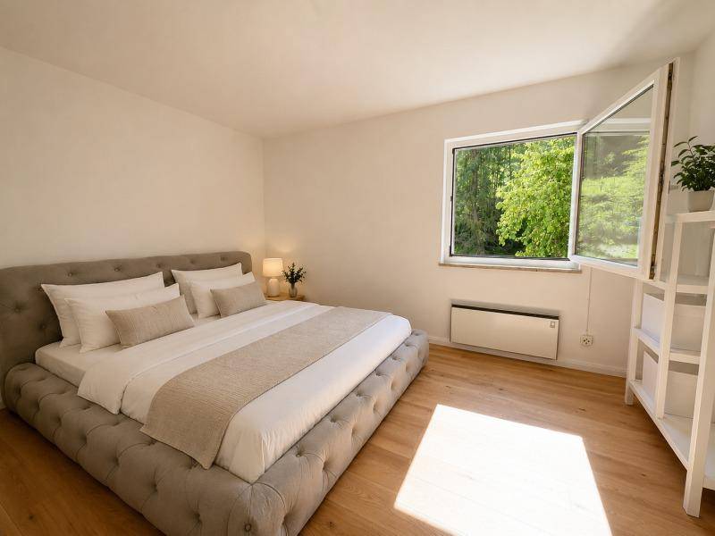 Helles Schlafzimmer mit großem Bett, Holzboden und Fenster mit Blick auf die grüne Natur.