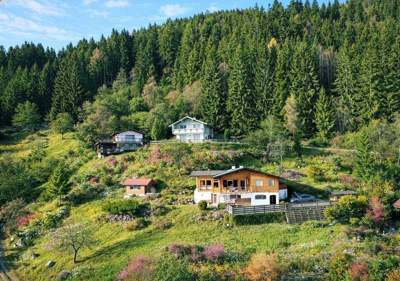 Mehrere Häuser in einer grünen Hügellandschaft, umgeben von Wäldern und blühenden Sträuchern im Sommer.
