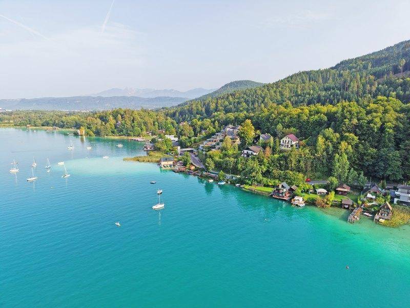 Luftaufnahme des Sees mit klarem Wasser, umgeben von üppiger Vegetation und vereinzelten Häusern.