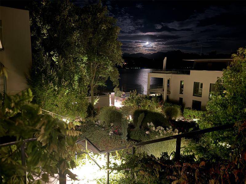 Beleuchteter Gartenbereich bei Nacht mit Blick auf den See und den Mond am Himmel.