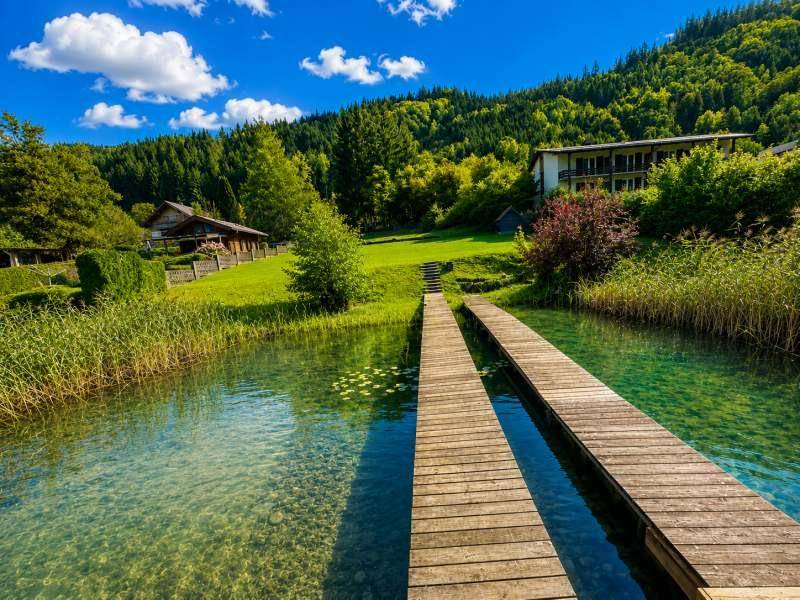 Blick vom Holzsteg über das klare Seewasser zu bewaldeten Hügeln und Gebäuden am Ufer.