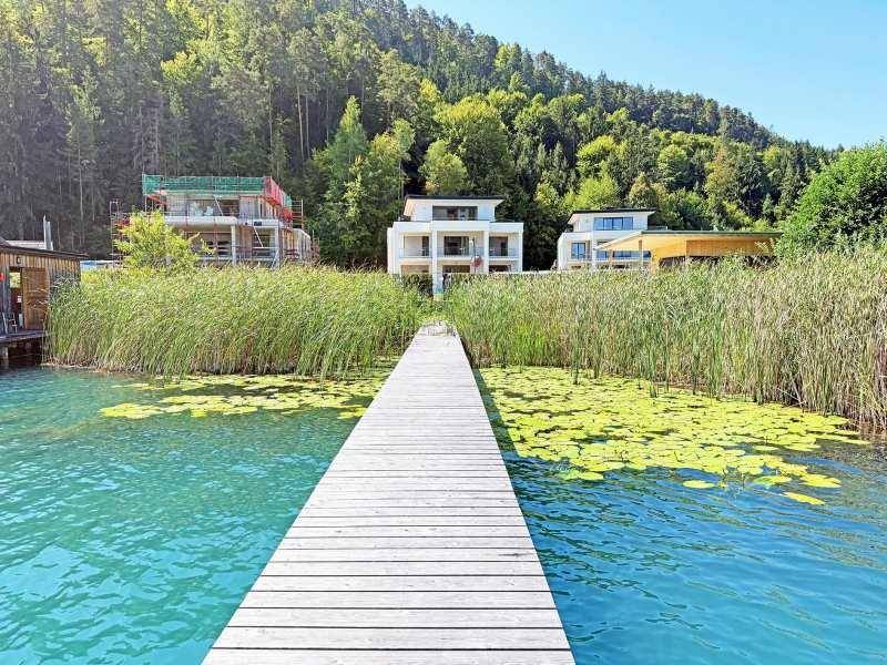 Moderner Holzsteg am Seeufer mit Blick auf zeitgenössische Häuser und bewaldete Hügel im Hintergrund.