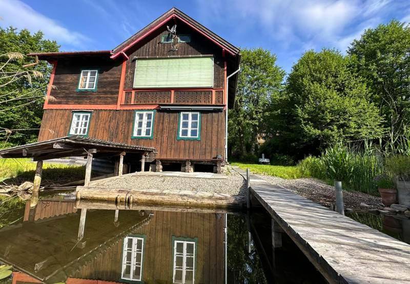 Das Holzhaus steht direkt am Wasser, mit einem Holzsteg, der zum See führt, umgeben von üppigem Grün.