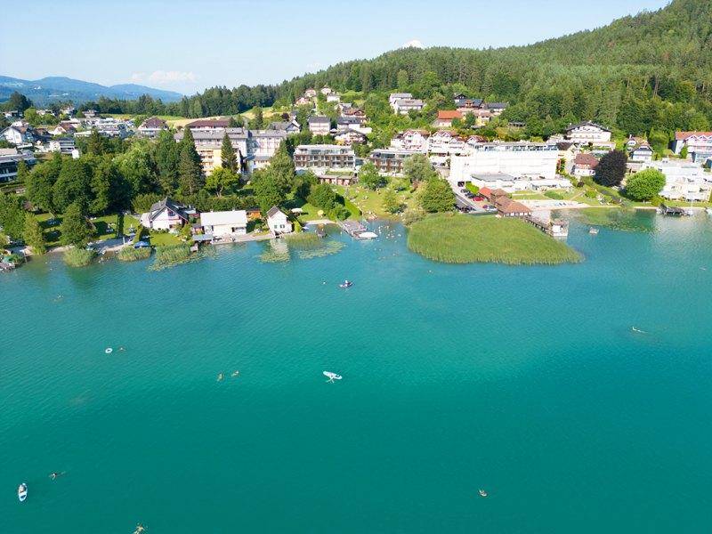 Luftaufnahme des lebhaften Seeufers mit Badegästen, Booten und umliegenden Häusern an einem sonnigen Tag.