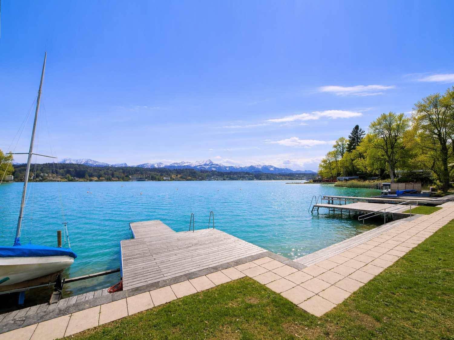 Panoramablick auf den See mit Holzstegen, einem Segelboot und schneebedeckten Bergen im Hintergrund.