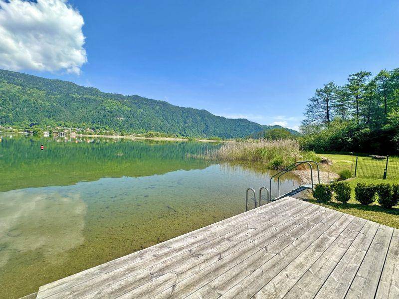 Holzsteg führt ins klare Seewasser mit Bergblick und Schilf am Ufer.