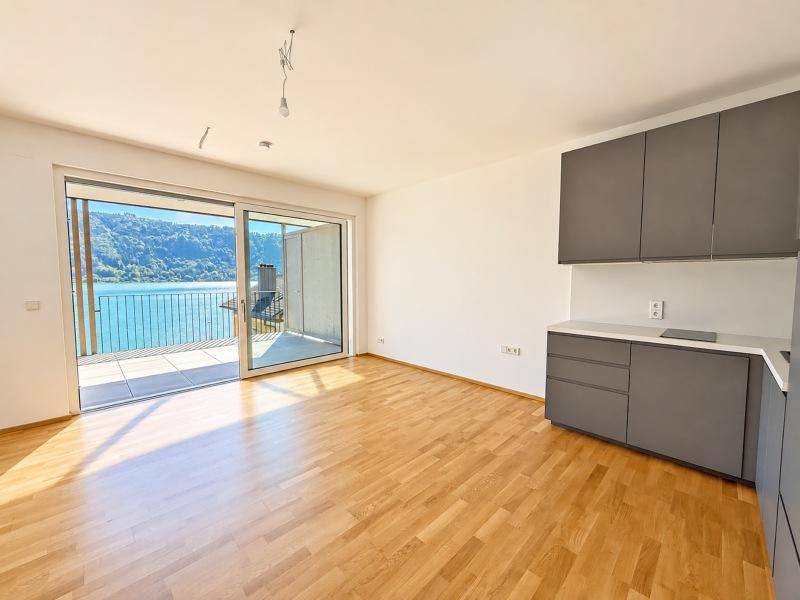 Heller Wohnbereich mit Holzboden, kleiner Küchenzeile und Zugang zu einem Balkon mit Seeblick.
