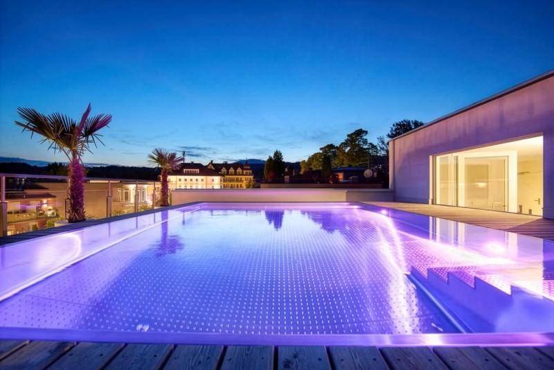 Beleuchteter Infinity-Pool auf einer Dachterrasse bei Sonnenuntergang mit Palmen.