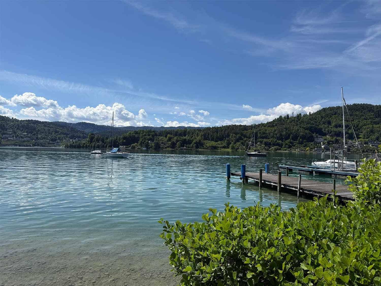 Klarer See mit Holzsteg und Segelbooten, umgeben von grünen Hügeln unter blauem Himmel.