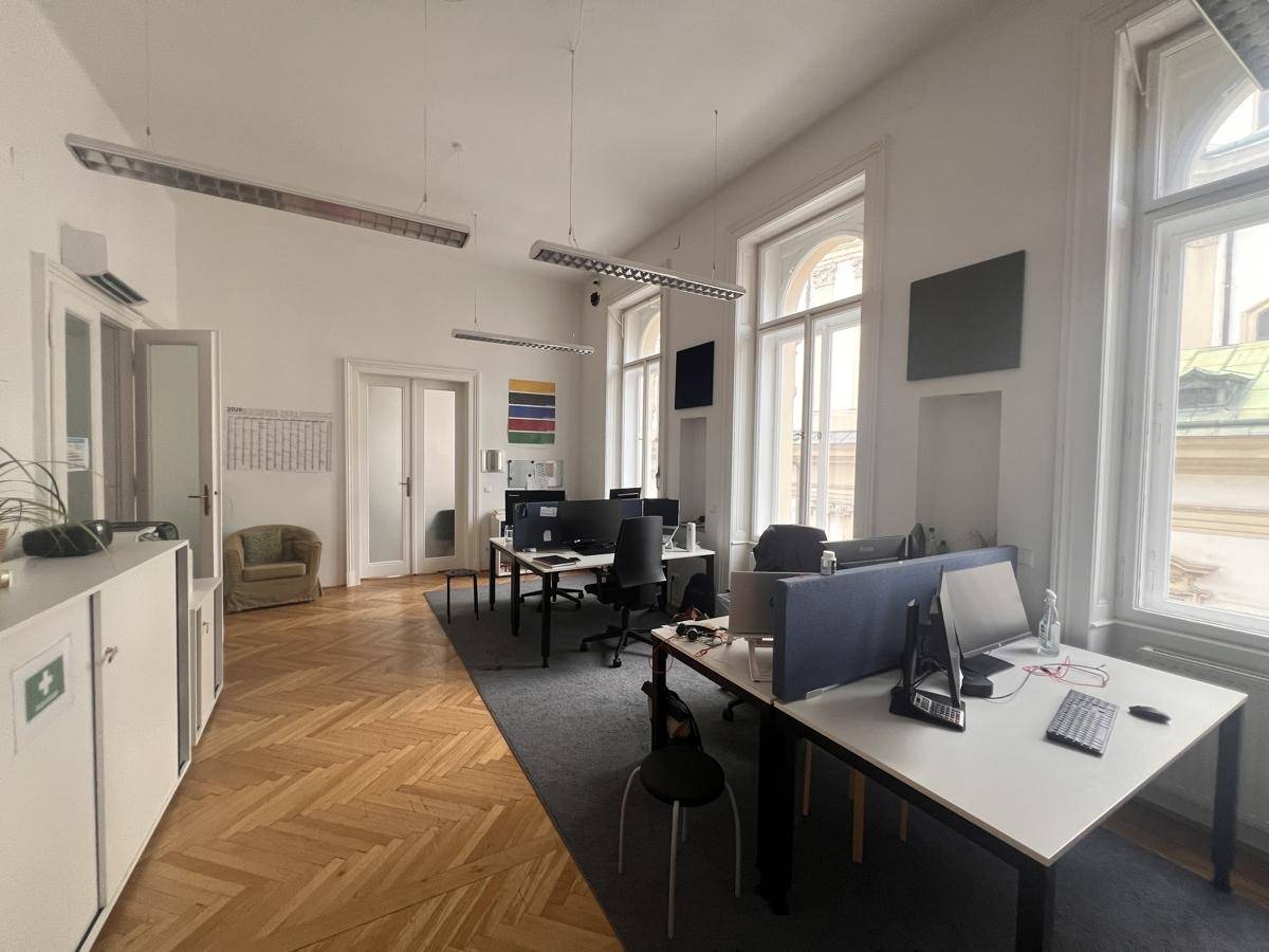 Großes offenes Büro mit mehreren Arbeitsplätzen, Fischgrätparkett und hohen Fenstern, gut beleuchtet.