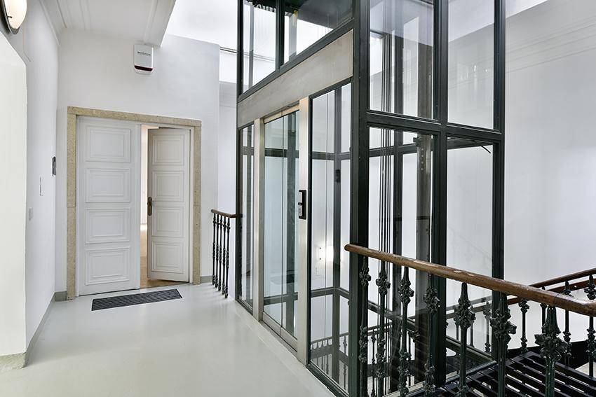 Heller Flur mit modernem Glasaufzug und klassischer Holztreppe im Gebäude.