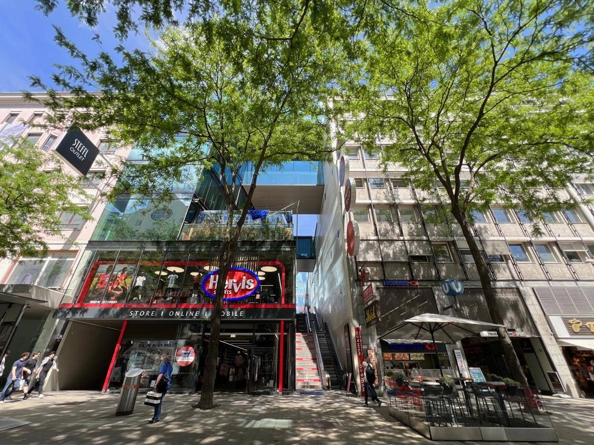 Eine belebte Einkaufsstraße mit modernen Gebäuden, Geschäften und Bäumen unter blauem Himmel.