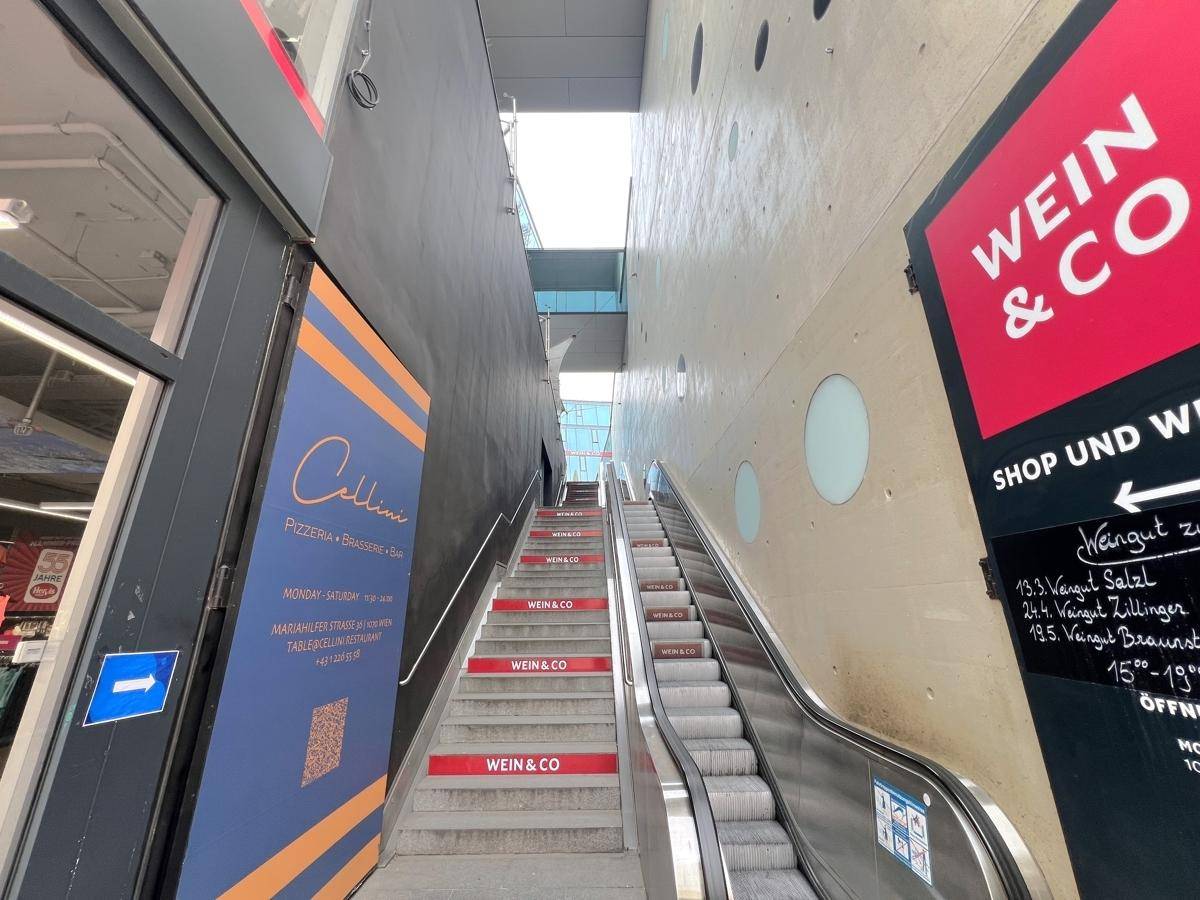 Eine Rolltreppe und Treppen führen zwischen zwei Gebäuden hinauf, flankiert von Werbeschildern.