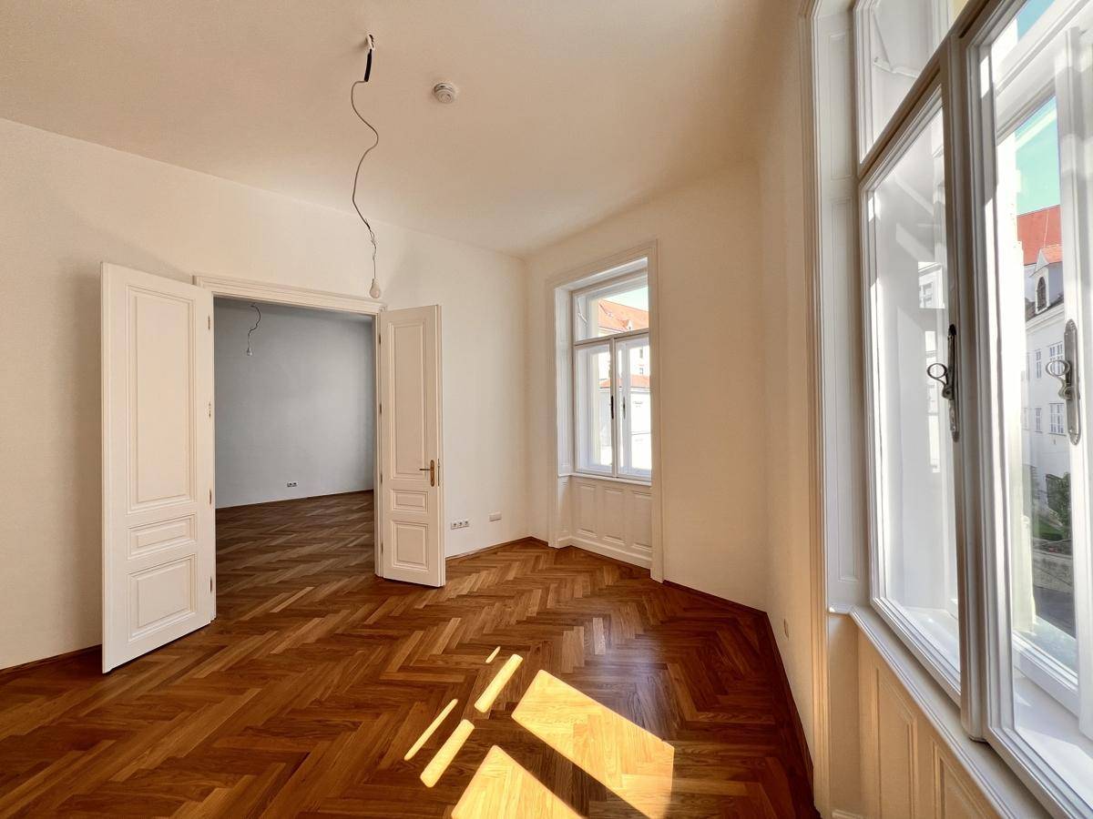 Lichtdurchfluteter Raum mit klassischem Fischgrätparkett und Zugang zu einem weiteren Zimmer.