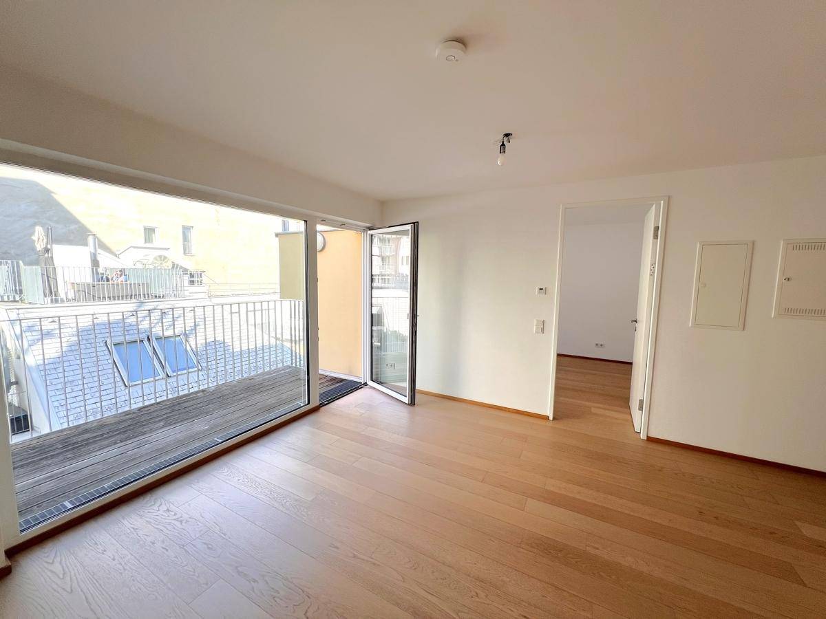 Geräumiger, leerer Wohnbereich mit hellem Holzboden und großen Fenstern zum Balkon.