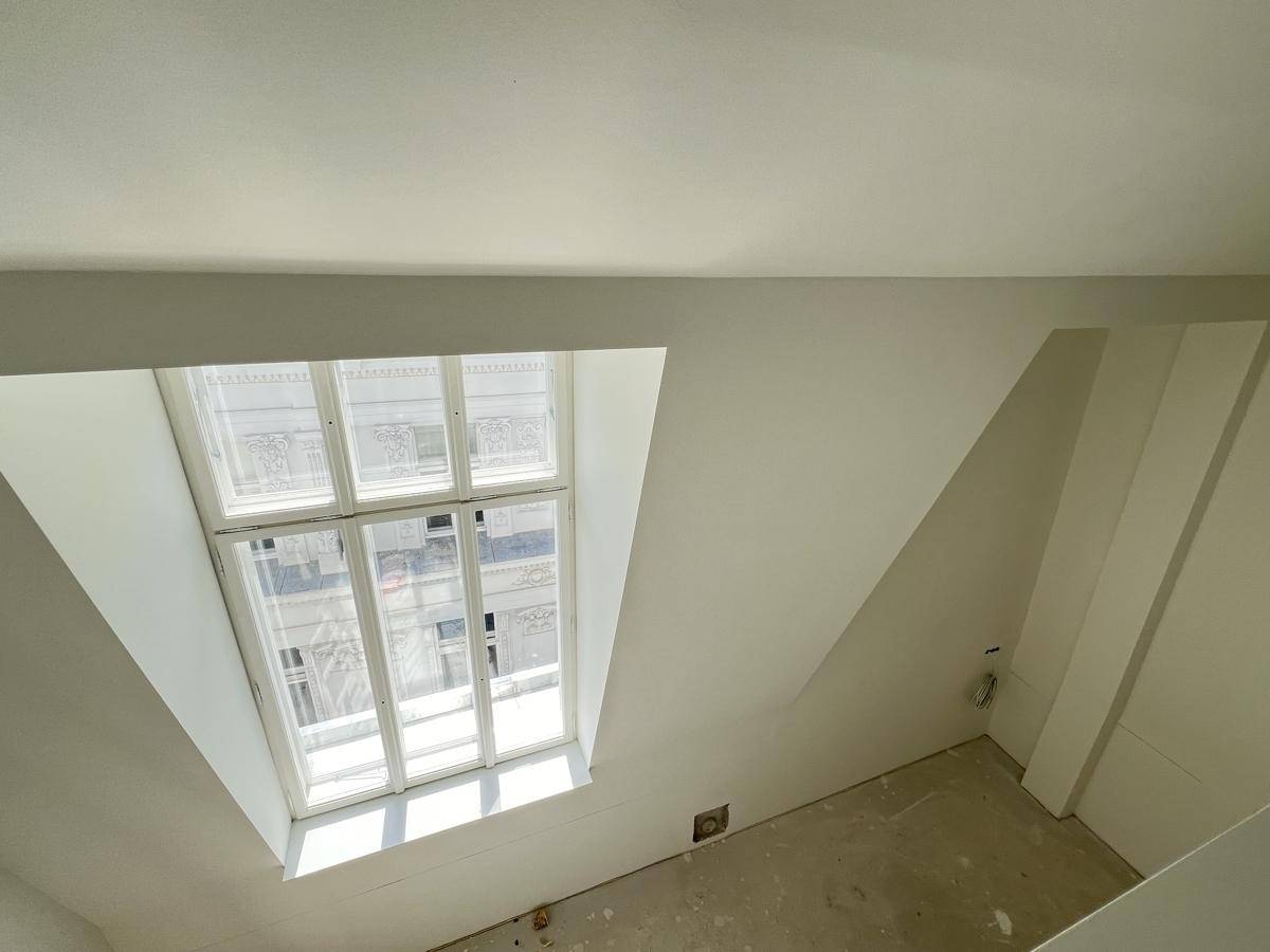 Dachgeschossraum im Rohbau mit schrägen Wänden und einem Fenster, das auf die Stadt blickt.