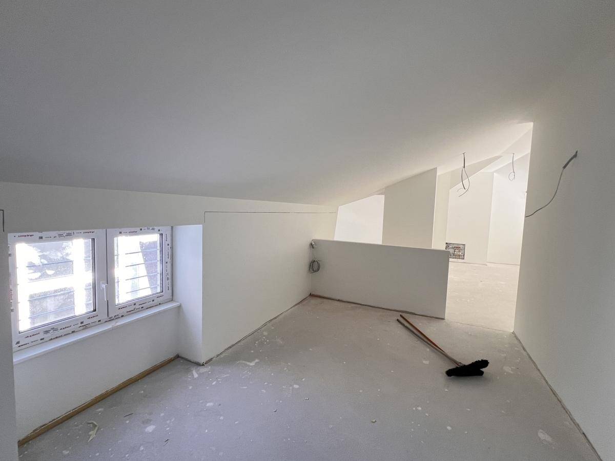 Unfertiger Dachgeschossraum mit schrägen Wänden, kleinem Fenster und Betonboden, bereit zum Ausbau.