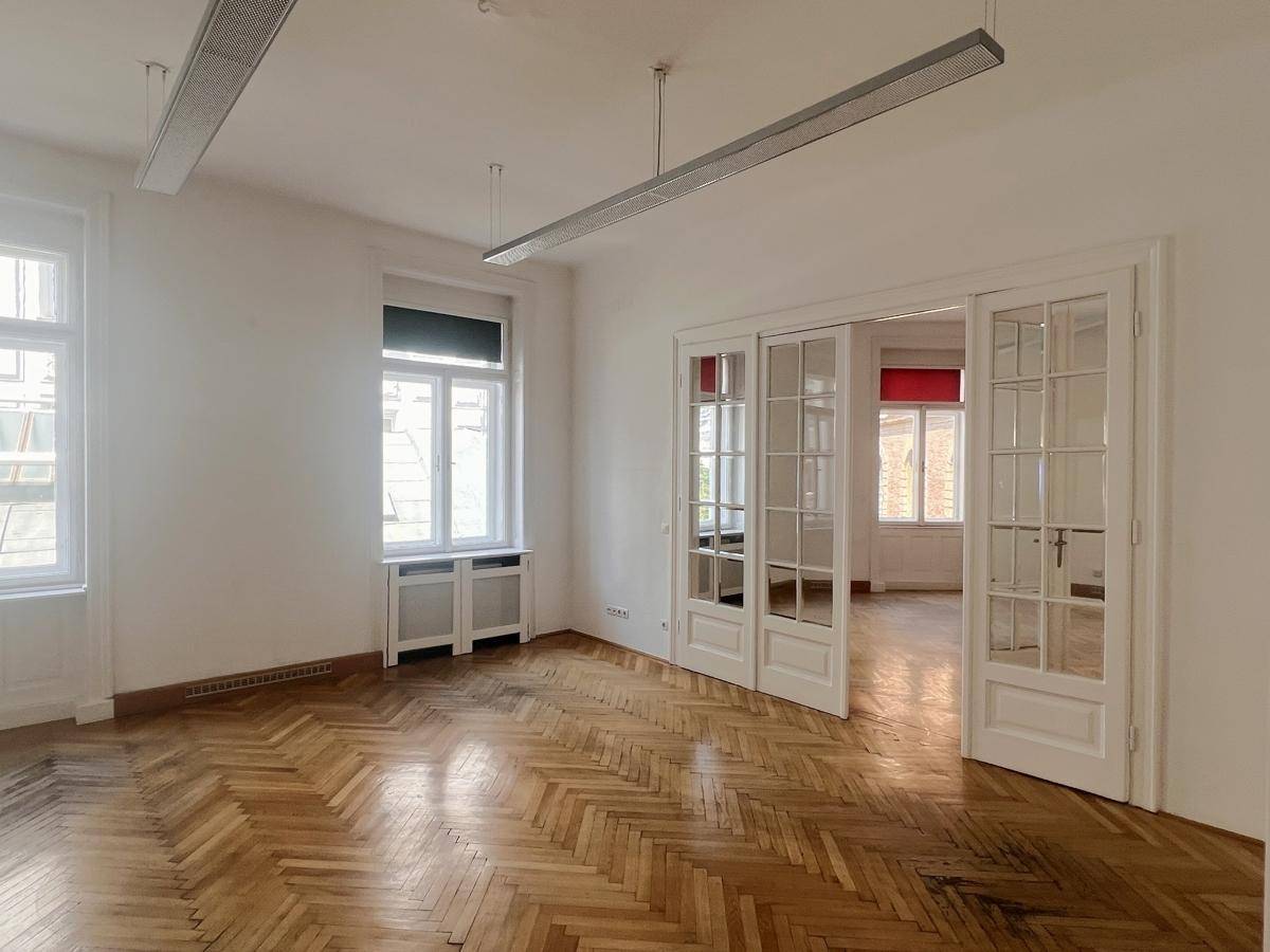 Geräumiges Büro mit Fischgrätparkett, hohen Fenstern und eleganten Glastüren zu einem weiteren Raum.