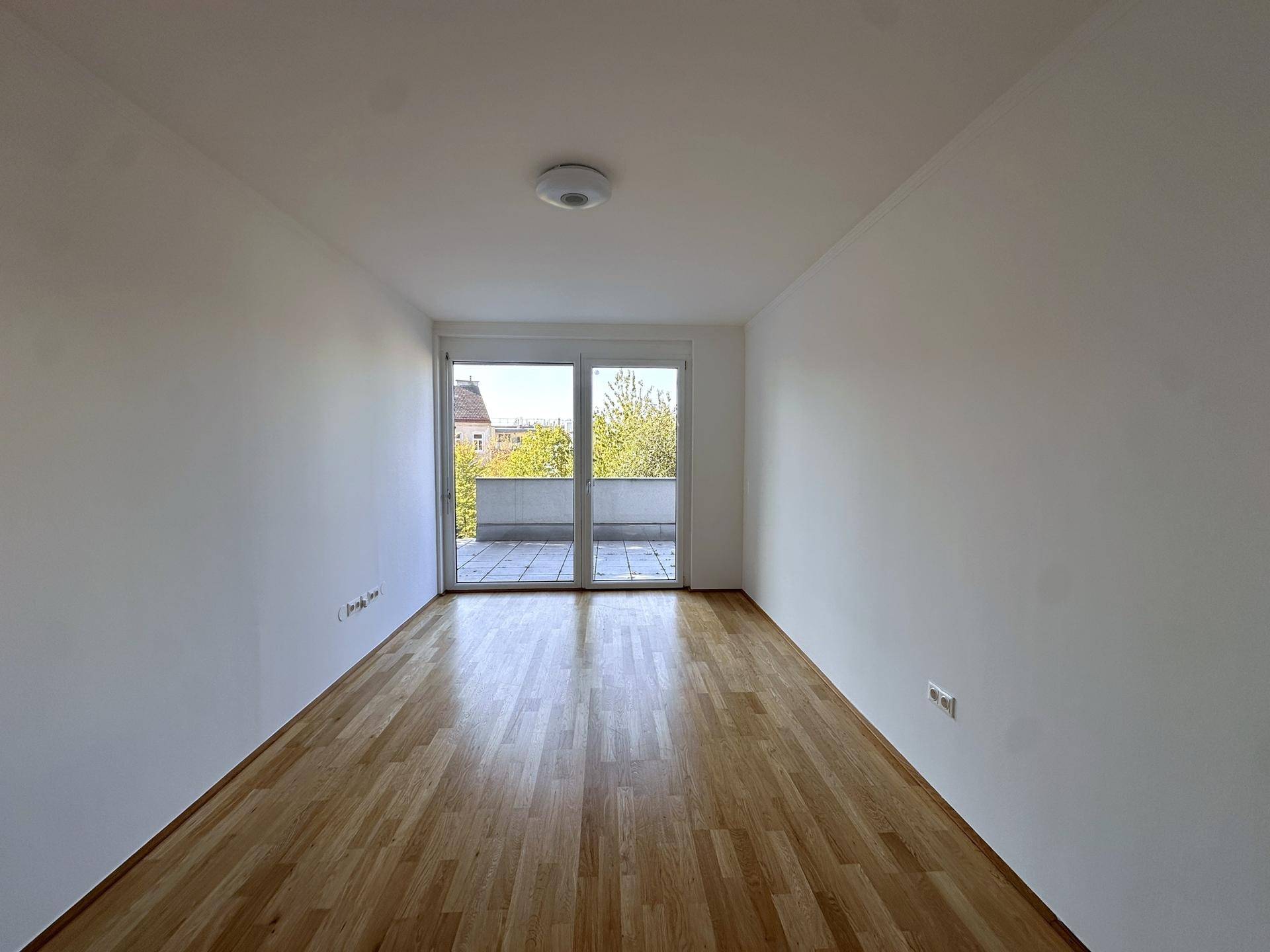 Helles, leeres Zimmer mit Holzboden und großen Fenstern zum Balkon, viel Tageslicht.