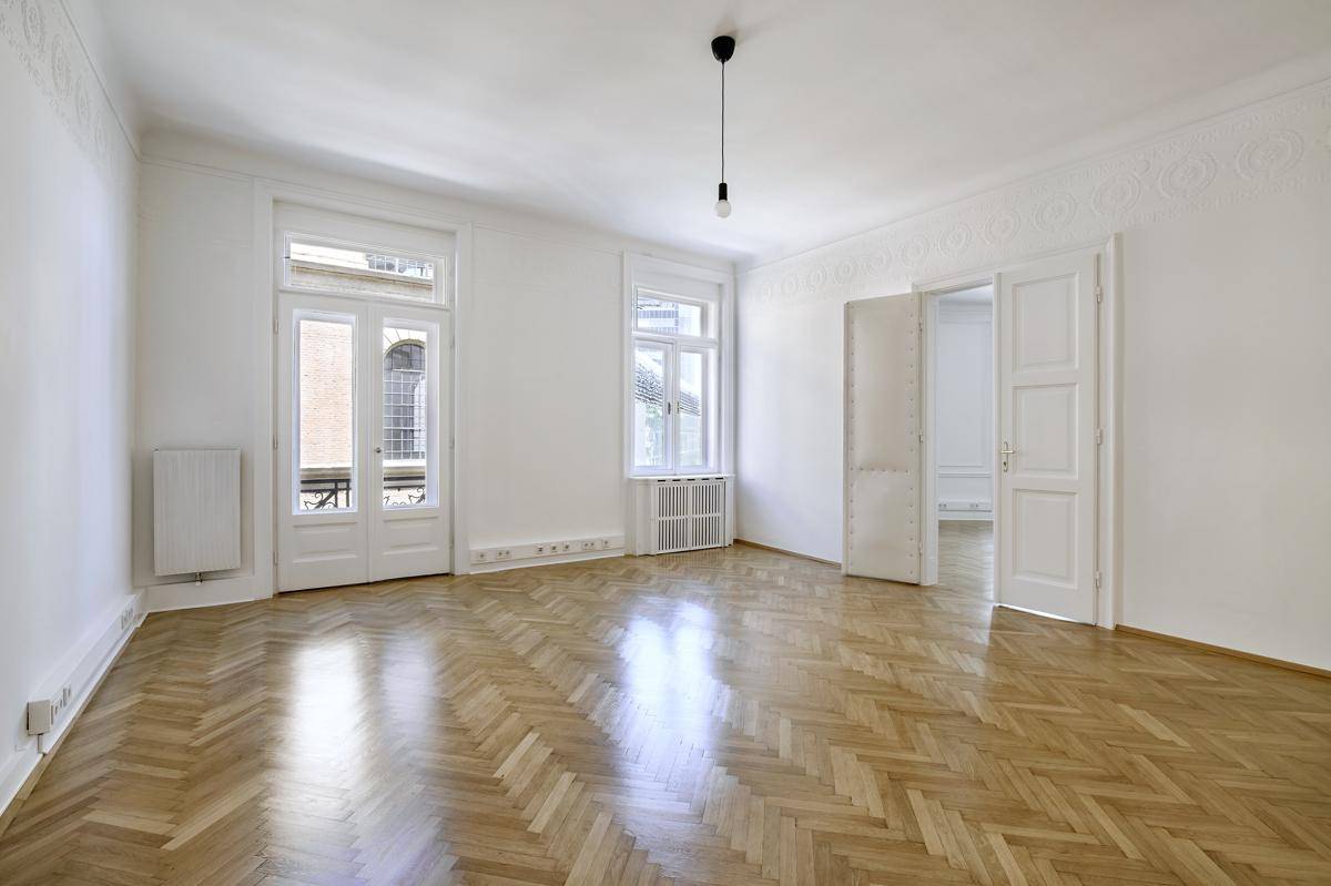 Heller Raum mit Fischgrätparkett, hohen Fenstern und klassischer Stuckverzierung an der Decke.