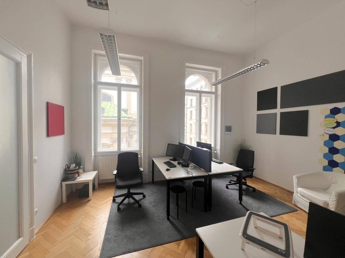 Helles Einzelbüro mit zwei Arbeitsplätzen, hohen Fenstern und Fischgrätparkett, schlicht eingerichtet.