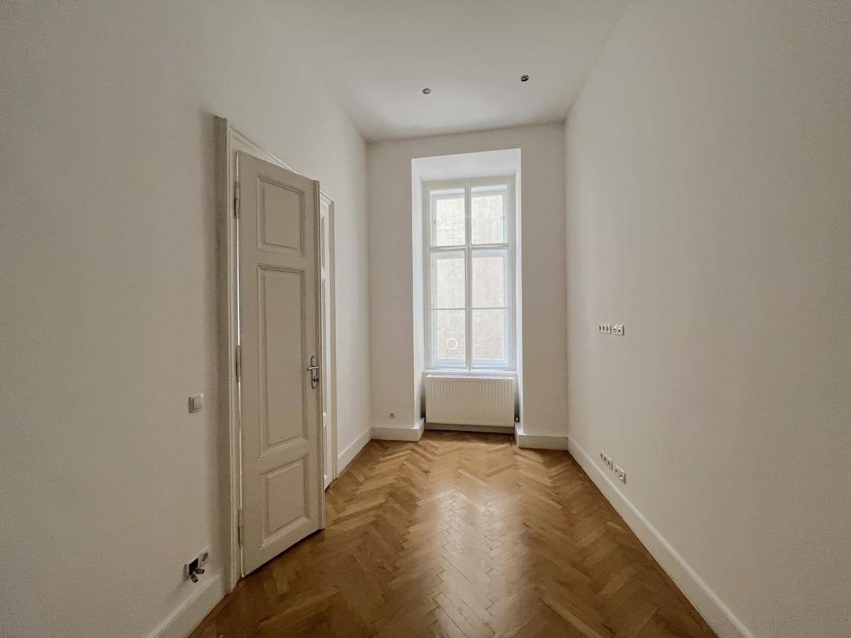 Schmaler Raum mit Fischgrätparkett, einem Fenster und einer offenen Tür.