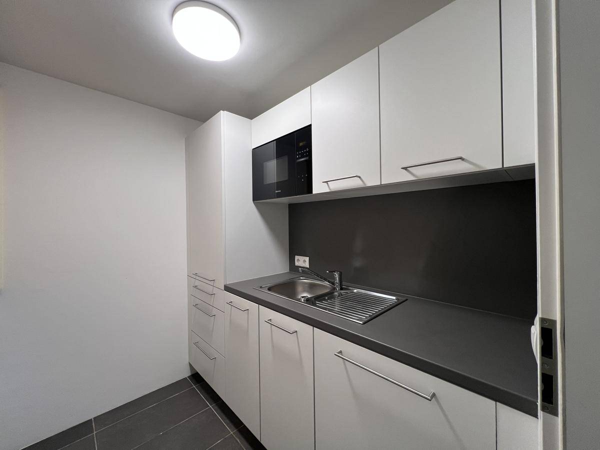 Moderne, kompakte Küchenzeile mit weißen Schränken, Spüle und integrierter Mikrowelle, ideal für kleine Büros.