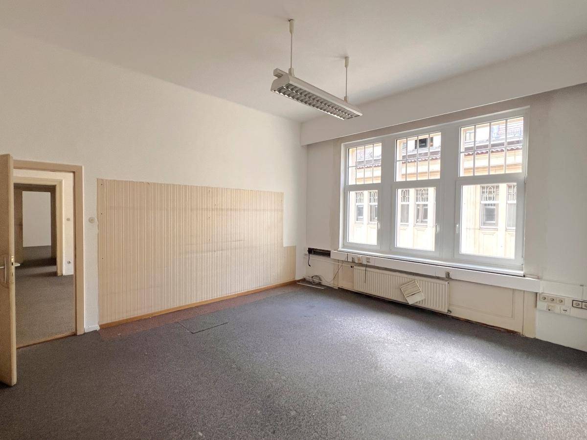 Leeres Büro mit dunklem Teppichboden, weißen Wänden und einem großen Fenster, das viel Tageslicht hereinlässt.