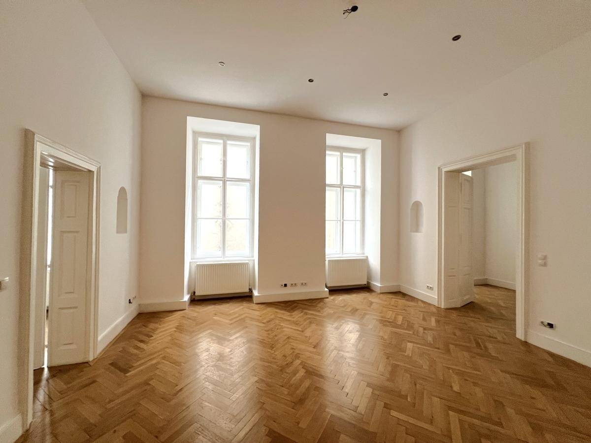 Geräumiges Zimmer mit klassischem Fischgrätparkett und viel Tageslicht durch hohe Fenster.
