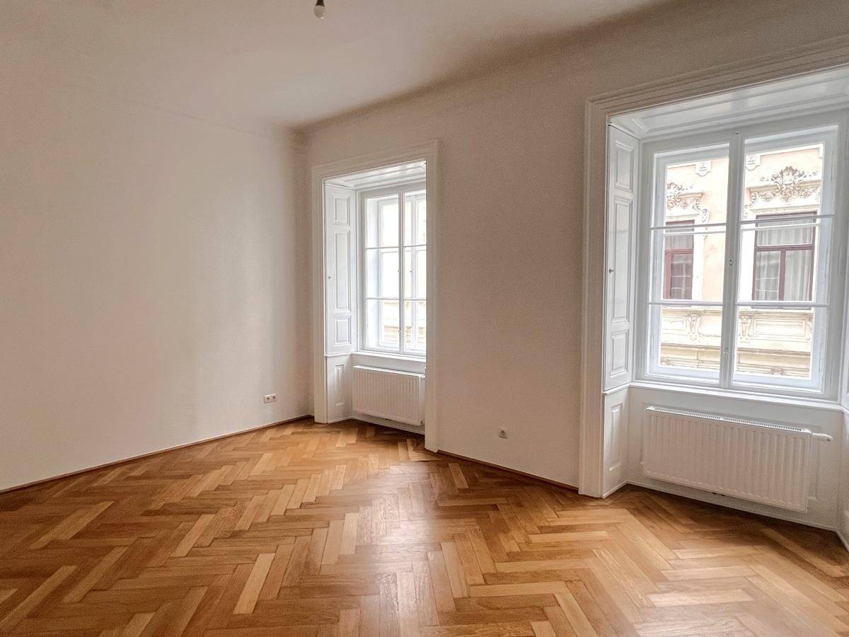 Geräumiges, leeres Zimmer mit Fischgrätparkett, hohen Fenstern und viel Tageslicht.