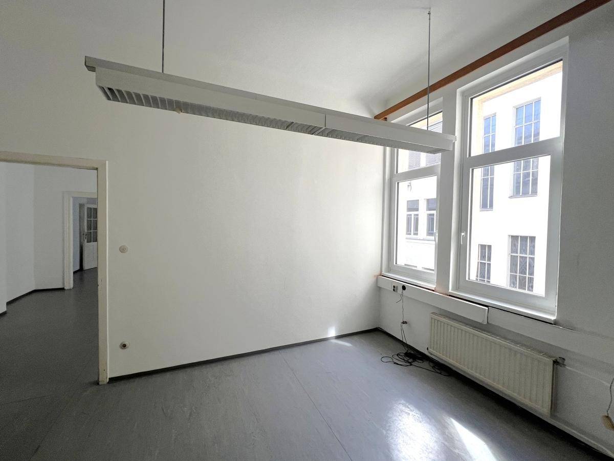 Helles Büro mit grauem Boden, weißen Wänden und zwei großen Fenstern, die viel Tageslicht hereinlassen.
