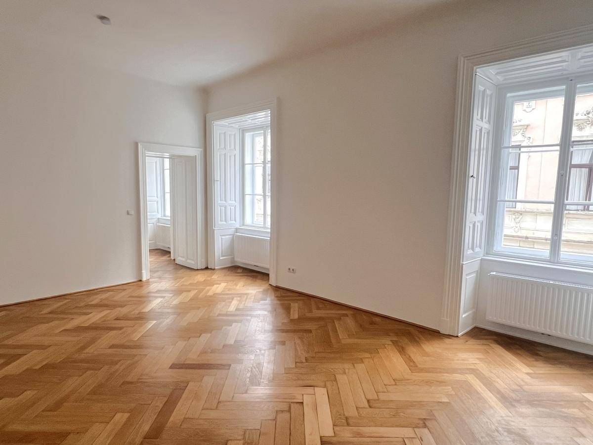 Geräumiges, leeres Zimmer mit Fischgrätparkett, hohen Fenstern und viel Tageslicht.