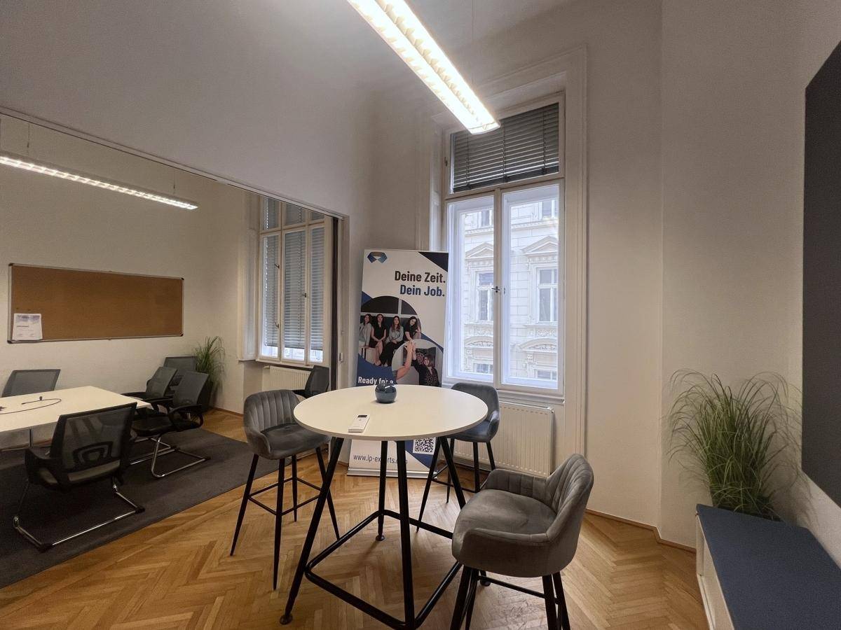 Flexibler Bürobereich mit Stehtisch und Stühlen, großen Fenstern und Parkettboden, hell und offen.
