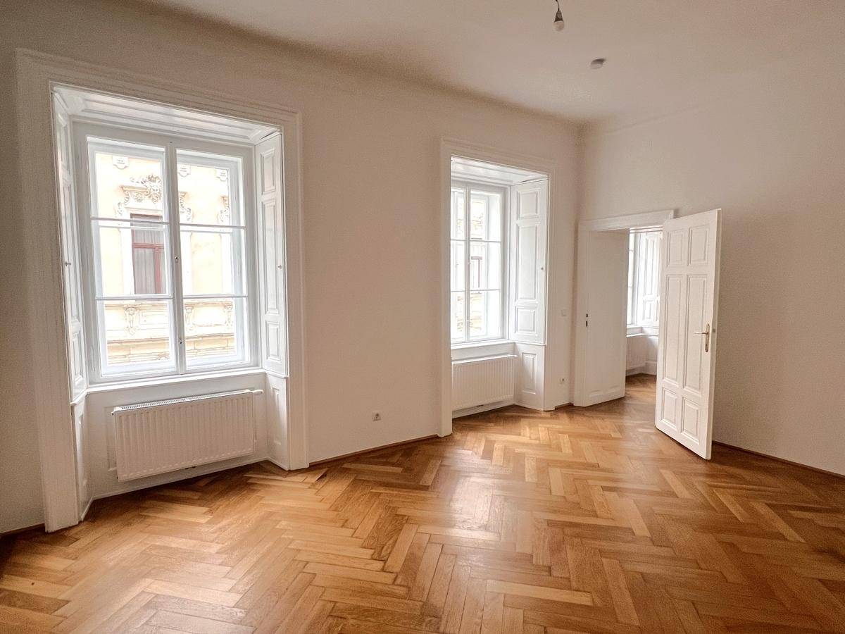 Geräumiges, leeres Zimmer mit Fischgrätparkett, hohen Fenstern und viel Tageslicht.