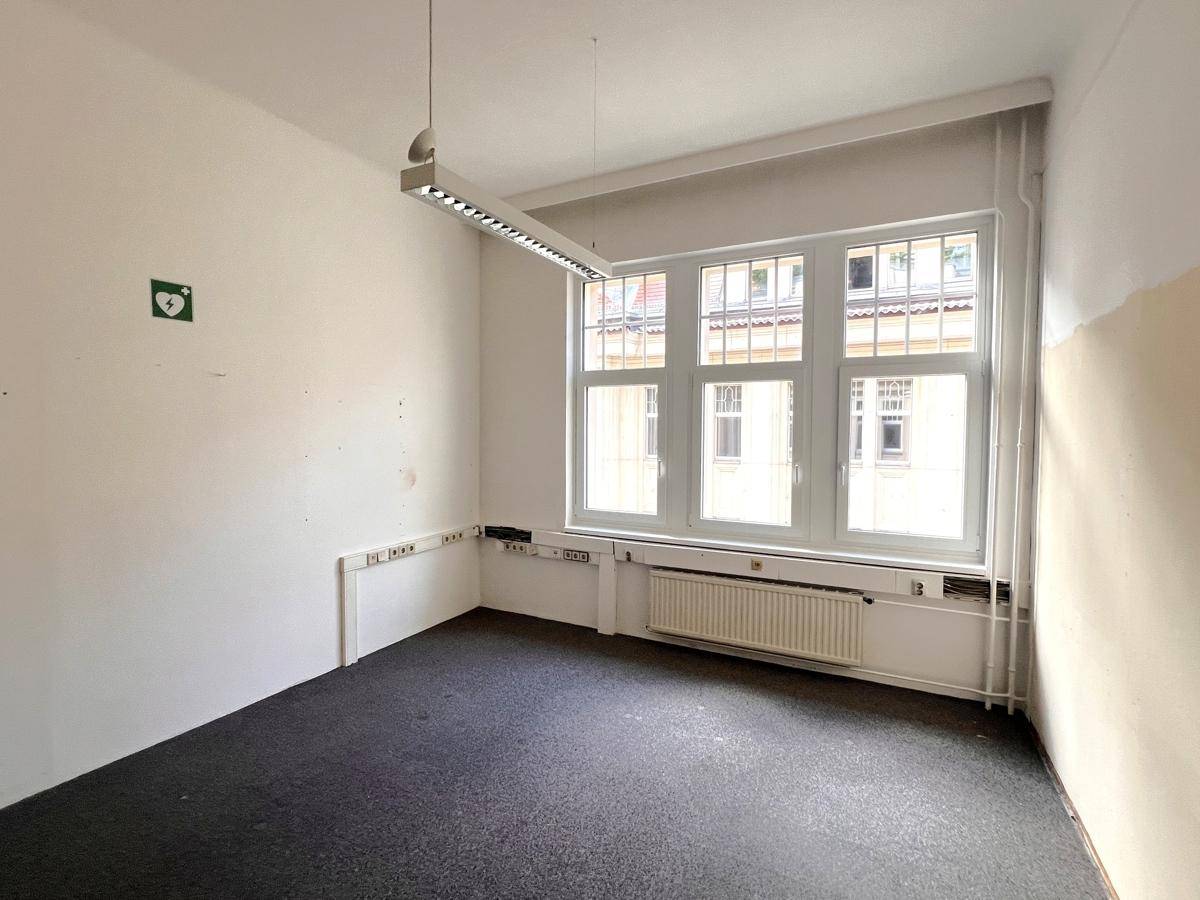 Helles, leeres Büro mit dunklem Teppichboden, großen Fenstern und einem Defibrillator-Hinweis an der Wand.
