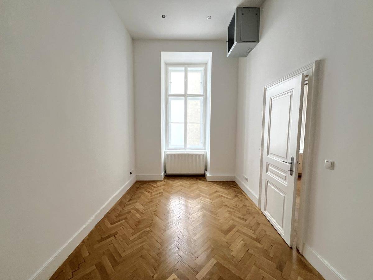 Schmaler Raum mit Fischgrätparkett, einem Fenster und einer Lüftungsanlage an der Decke.