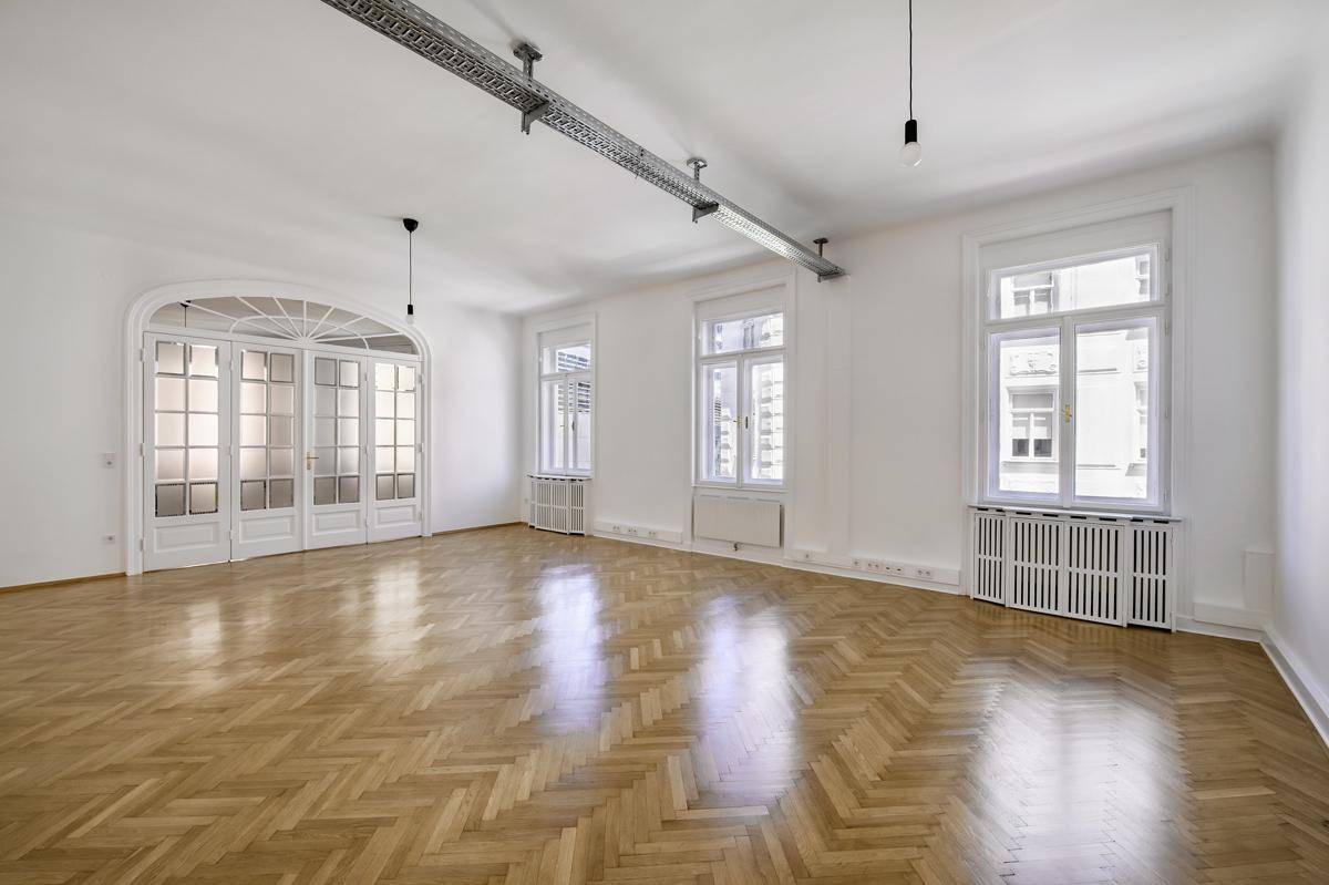 Heller Büroraum mit Fischgrätparkett, hohen Fenstern und sichtbaren Kabelkanälen an der Decke.