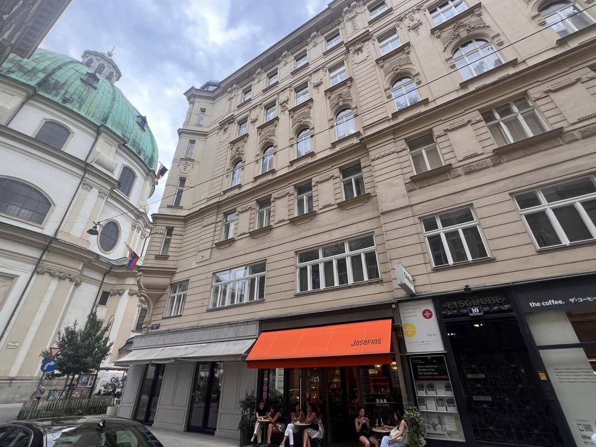 Historisches Gebäude an einer belebten Straße mit Geschäften im Erdgeschoss, unter bewölktem Himmel.