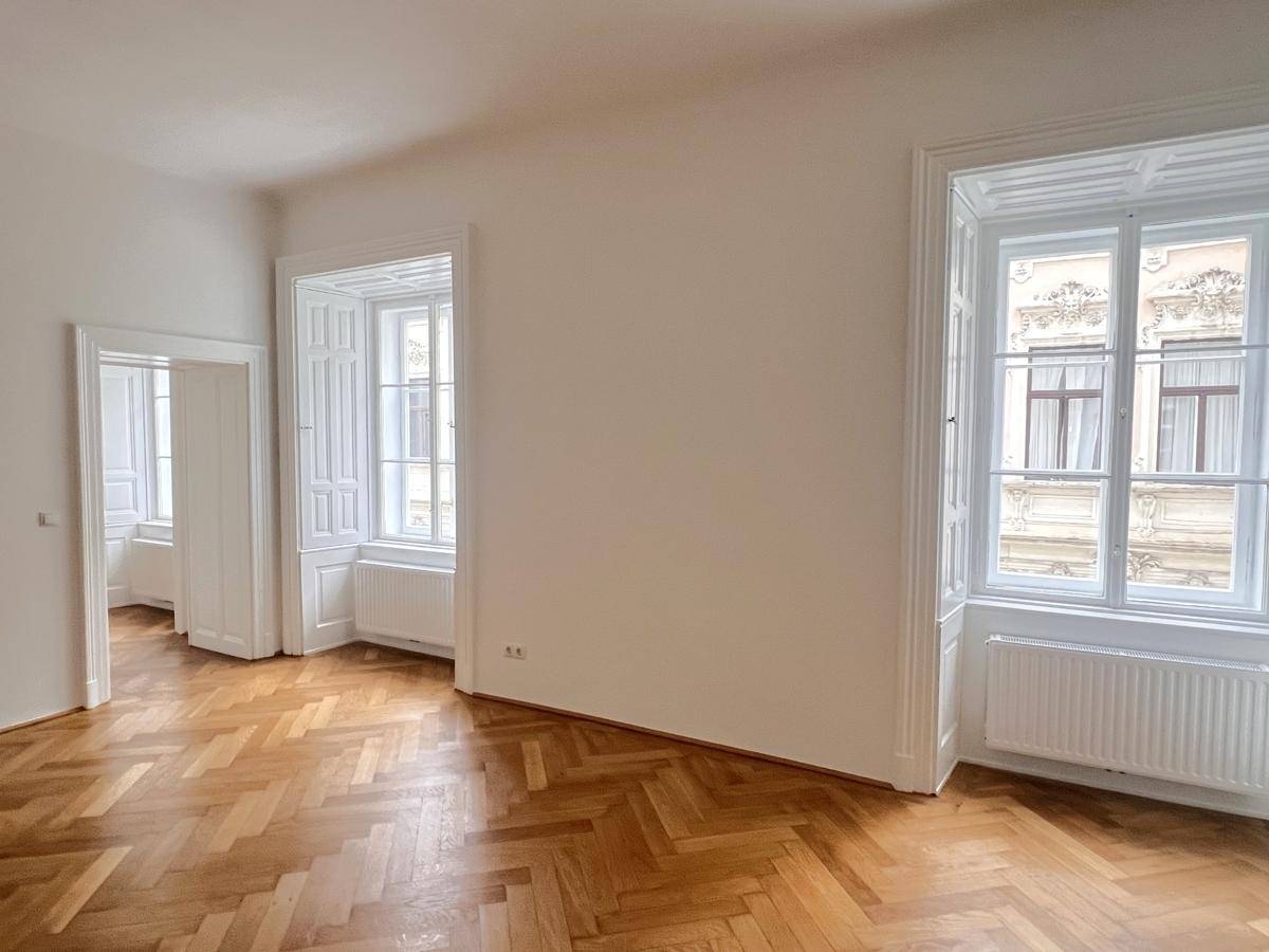 Geräumiges, leeres Zimmer mit Fischgrätparkett, hohen Fenstern und viel Tageslicht.