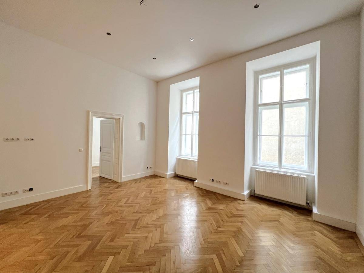 Großzügiges Zimmer mit klassischem Fischgrätparkett, hohen Fenstern und weiß gestrichenen Wänden.