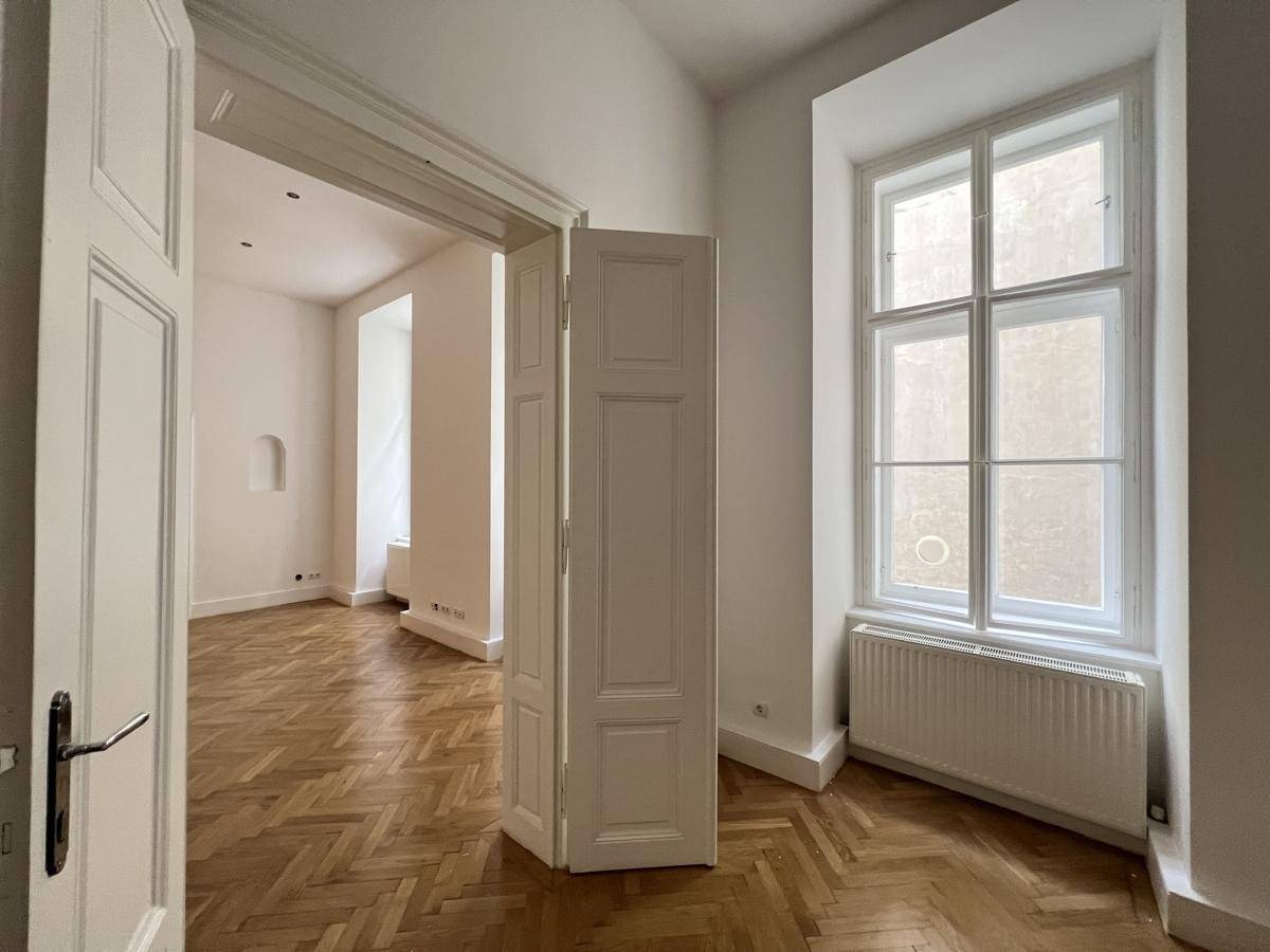 Blick durch eine offene Tür in ein helles Zimmer mit Fischgrätparkett und hohen Fenstern.