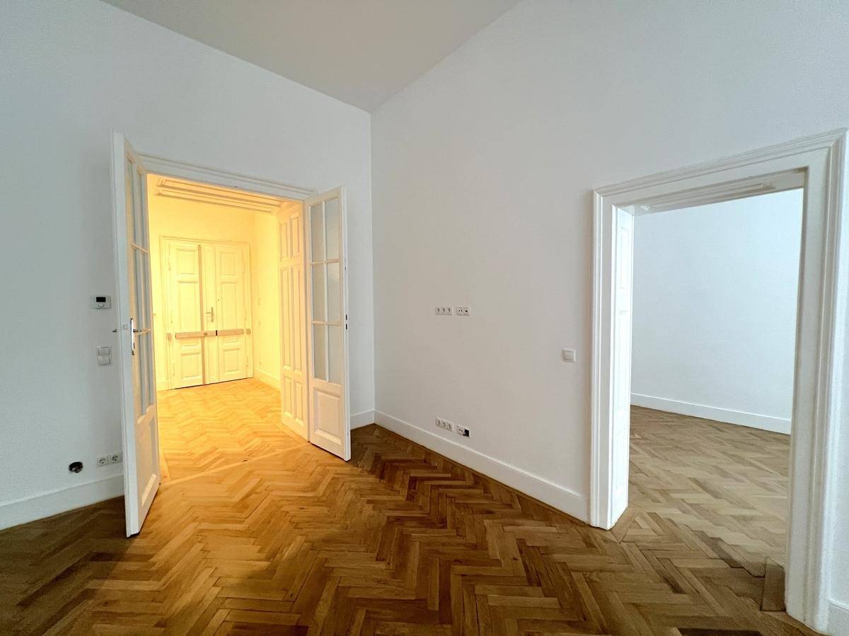 Heller Raum mit Fischgrätparkett und eleganten Flügeltüren, die zu einem weiteren Zimmer führen.