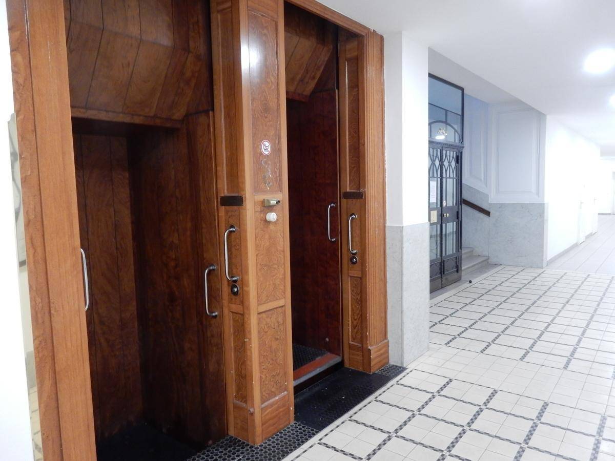 Historischer Paternoster-Aufzug aus Holz in einem eleganten Flur mit gemusterten Fliesen und Marmorwänden.